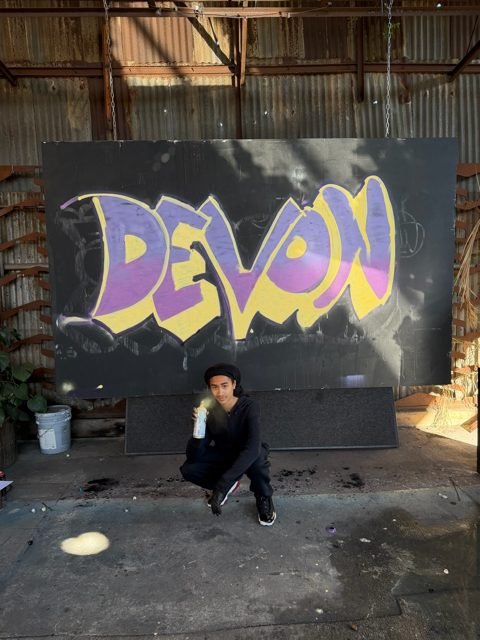 Workshop guest posing beside a purple and yellow DEVON graffiti piece
